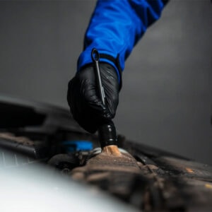 Maintenance worker cleaning car engine with brush, wearing blue uniform and black gloves.